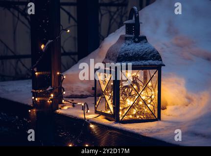 Schwarze Metall-Kerzenlaterne mit LED-Lichtern, die im Winter draußen drinnen leuchten. Verschneite Holzterrasse in der Nacht, während der Weihnachtsferien. Stockfoto