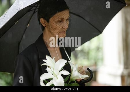 Seitenansicht einer älteren Frau unter schwarzem Schirm mit Trauerschleier und trauerndem Verlust eines verstorbenen Angehörigen am Grab, während sie weiße Lilien auf dem Friedhof hält, Kopierraum Stockfoto