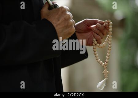 Nahaufnahme der Hände einer nicht erkennbaren Seniorin, die hölzerne Rosenkranzperlen hält und für die Seele des Verstorbenen betet, während sie sich vor Regen unter dem Schirm versteckt, Kopierraum Stockfoto