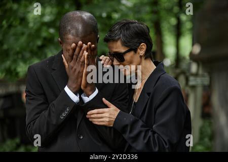 Mittlere Aufnahme einer älteren Frau in Sonnenbrille, die den schreienden Ehemann bei der Gedenkfeier tröstet. Multiethnisches Paar trauert den Verlust eines geliebten Menschen auf dem Friedhof Stockfoto