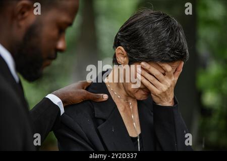Mittlere Aufnahme einer älteren Frau in schwarzem Anzug, die das Gesicht mit der Hand bedeckt, die über den Verlust eines geliebten Menschen trauert, und eines schwarzen Mannes, der seine Frau bei der Gedenkfeier am Grabstein tröstet Stockfoto