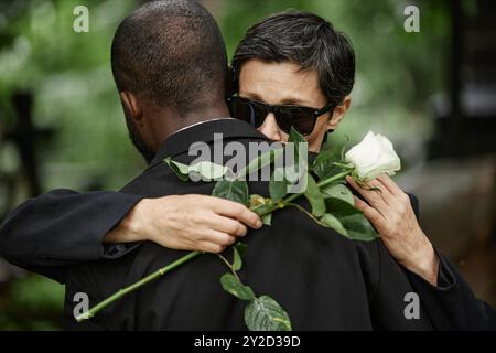 Mittelgroßer Schuss einer älteren Frau mit weißer Rose in der Hand, die einem nicht erkennbaren schwarzen Mann tröstende Umarmung gibt, der über den Verlust eines geliebten Menschen auf dem Friedhof trauert Stockfoto