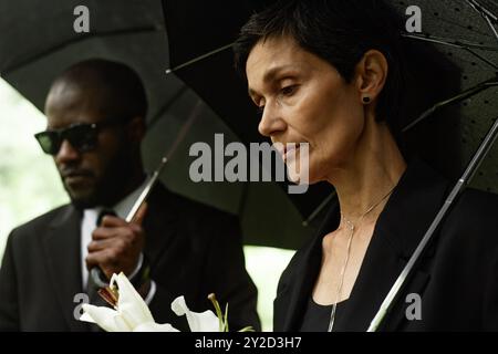 Seitenansicht einer traurigen Seniorin, die auf den schweren Verlust eines geliebten Menschen herabblickt, während sie während der Gedenkfeier auf dem Friedhof unter einem Schirm steht, Kopierraum Stockfoto