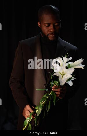 Vertikale Aufnahme des nachdenklichen Schwarzen Mannes mit Lichtstrahl, der auf elegantem Anzug leuchtet, der zarte weiße Lilie hält und im Studio mit dimmem Blitz vor schwarzem Hintergrund posiert Stockfoto