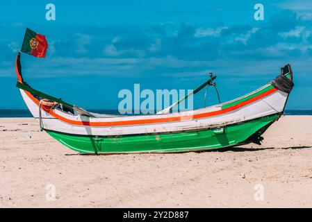 Am Sandstrand ruht ein hell bemaltes Moliceiro-Boot mit portugiesischer Flagge Stockfoto