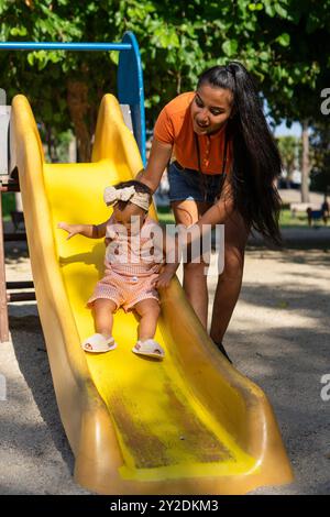 Lateinische Mutter mit ihrer einjährigen Tochter auf der Rutsche eines Spielplatzes Stockfoto