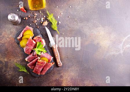 Getrocknete Pastrami-Scheiben, Rindfleisch mit Kräutern und eingelegte Gurke auf schwarzer Platte. Stockfoto