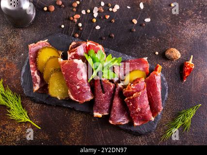 Getrocknete Pastrami-Scheiben, Rindfleisch mit Kräutern und eingelegte Gurke auf schwarzer Platte. Stockfoto