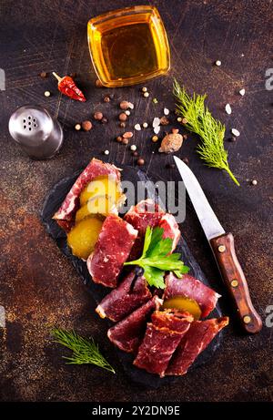 Getrocknete Pastrami-Scheiben, Rindfleisch mit Kräutern und eingelegte Gurke auf schwarzer Platte. Stockfoto