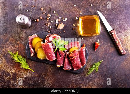 Getrocknete Pastrami-Scheiben, Rindfleisch mit Kräutern und eingelegte Gurke auf schwarzer Platte. Stockfoto