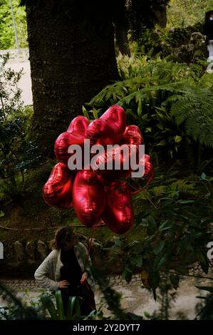 In Quinta da Regaleira schweben leuchtend rote Ballons in Herzform vor einer üppigen Gartenkulisse Stockfoto