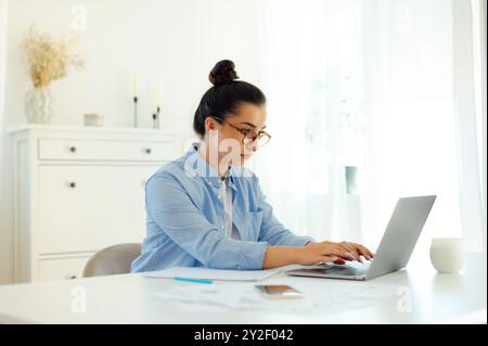 Fokussierte ernsthafte Geschäftsfrau arabischer oder indischer Herkunft in blauem Hemd, Digital Manager, ceo des Unternehmens, sitzt am Arbeitsplatz, während sie an einem Laptop arbeitet, konzentriert auf den Bildschirm schaut, nach Informationen sucht Stockfoto