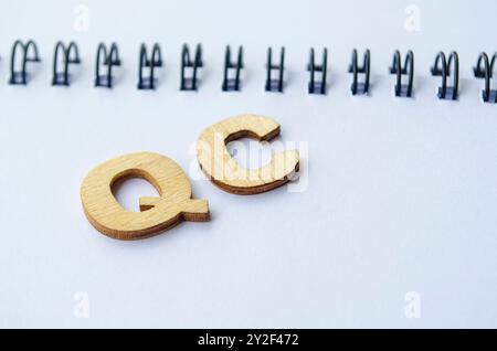 Draufsicht der QC-Holzbuchstaben auf weißem Notizblock. QK für Qualitätskontrolle. Stockfoto