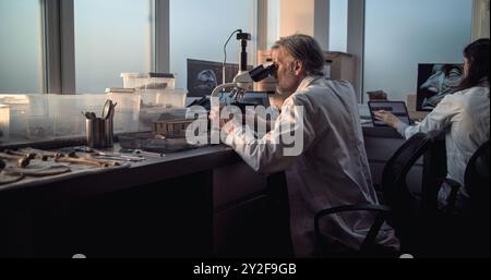 Ein ausgereifter Mikrobiologe führt Fossilienanalysen unter Mikroskop durch, schaut auf den Laptop-Bildschirm. Die Wissenschaftlerin arbeitet am PC und untersucht dann alte menschliche oder tierische Knochen. Fortgeschrittenes archäologisches Labor. Stockfoto