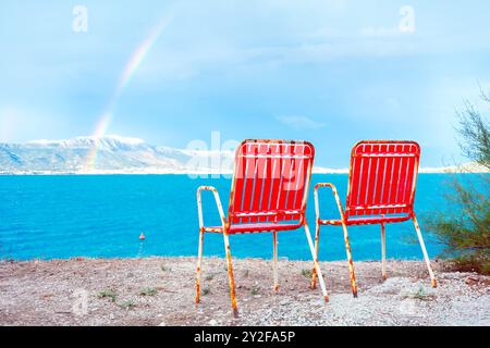 Rote Liegestühle stehen am Strand neben dem Meer. Zwei Stühle an der Küste. Regenbogen über dem Meer Stockfoto