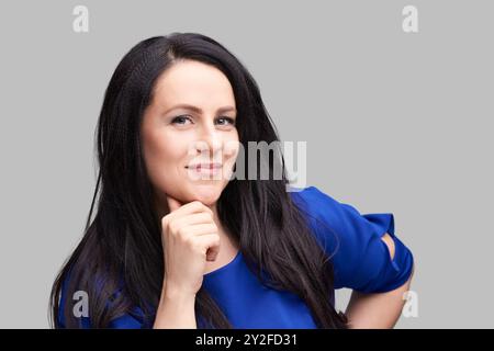 Selbstbewusste Frau in blauer Bluse mit langen schwarzen Haaren auf grauem Hintergrund. Stockfoto