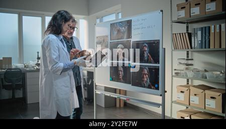 Männliche und weibliche Wissenschaftler untersuchen den fossilen Schädel des alten Mannes mit einem Tablet-Computer. Archäologen arbeiten im archäologischen Labor. Platine mit gedruckten Bildern zur 3D-Visualisierung der menschlichen Kopfanatomie. Stockfoto