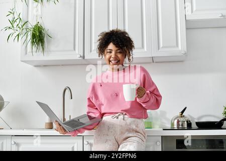 Eine junge Frau lächelt, während sie einen Becher und einen Laptop in ihrer hellen Küche hält. Stockfoto