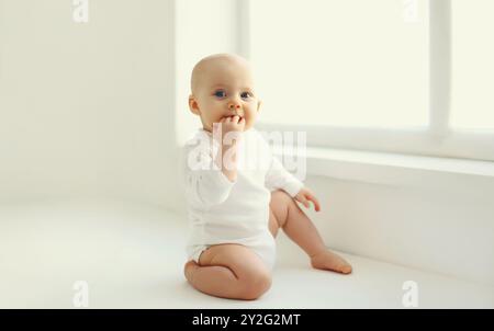 Niedliches kleines Baby, das auf dem Boden sitzt, beißt schmutzige Finger im weißen Zimmer zu Hause in der Nähe des Fensters Stockfoto