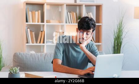 Online-Recherche. Fernunterricht. Internet-Bildung. Nachdenklicher, intelligenter Student mit Brille, der von zu Hause aus mit einem Laptop im modernen Innenraum des Arbeitsplatzes mit freiem Platz arbeitet. Stockfoto