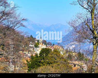 Der üppige Wald am Berghang öffnet den Blick auf die mittelalterliche Pfarrkirche Carona und die Ziegeldächer der Stadt gegenüber den trüben Alpen, Tessin, SW Stockfoto
