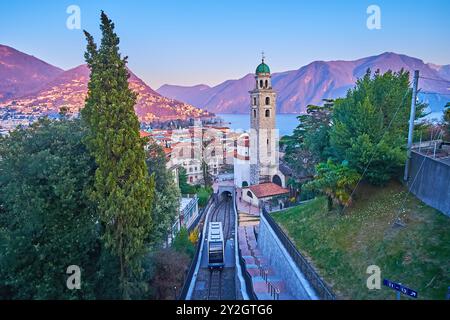 Blaue Stunde Lugano Stadtlandschaft mit Alpen und Ceresio See im lila Sonnenuntergang, hohem Glockenturm der Kathedrale San Lorenzo und Fahrwagen von Sassellina Fu Stockfoto