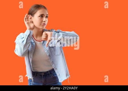 Ein Teenager-Mädchen, das versucht, etwas zu hören, und zeigt den Daumen nach unten auf orangem Hintergrund Stockfoto