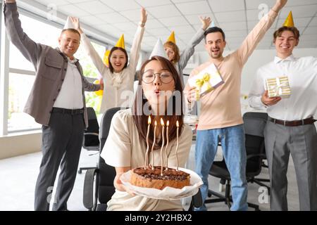 Junge asiatische Geschäftsfrau, die Kerzen auf Kuchen auf der Geburtstagsfeier im Büro ausbläst Stockfoto