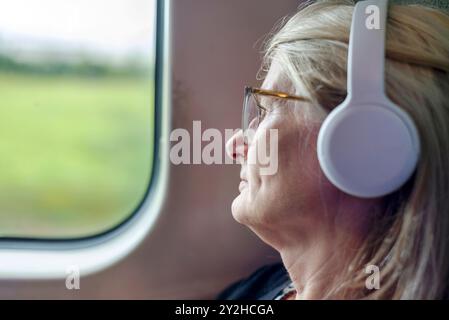 Eine blonde Frau, die in einem schnellen Fernzug reist, ein Hörbuch auf ihren großen Noise Cancelling-Ohrhörern hört, eine Brille trägt, passiv wat Stockfoto