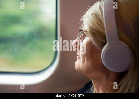 Eine blonde Frau, die in einem schnellen Fernzug reist, ein Hörbuch auf ihren großen Noise Cancelling-Ohrhörern hört, eine Brille trägt, passiv wat Stockfoto