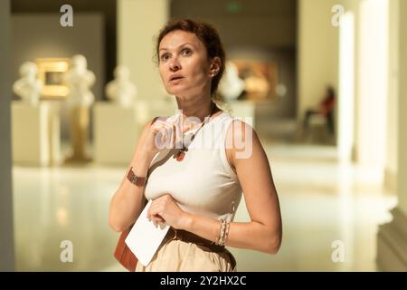 Interessierte Frau, die sich im Museum die klassischen Statuen genauer anschaut und den Tourbogen in der Hand hält Stockfoto