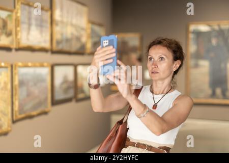 Interessierte Frau, die sich Gemälde in der Kunstgalerie anschaut, ihr Smartphone benutzt und Fotos macht Stockfoto