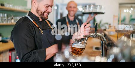 Stilvoller bärtiger Barkeeper in schwarzer Uniform, der Bier an der Bar anzapft und der Kellner mit Tablett plauderte miteinander und lächelte miteinander. Erfolgreiches People Team Wo Stockfoto