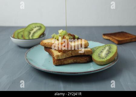 Sie gießt Honig, Sirup über ein Sandwich mit Brot, Tiost und Kiwis auf grauem Hintergrund. Das Konzept von gesunden Snacks und leckeren Speisen. Stockfoto