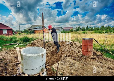 Der Baumeister platziert einen Betonring auf einen anderen und ebnet ihn mit einem Hebel mit einem Stahlrohr beim Bau einer Klärgrube. Stockfoto