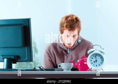 Ein junger Büroangestellter gähnt mit einer Tasse Tee und einem Wecker vor seinem Schreibtisch. Er ist erschöpft, nachdem er lange aufgeblieben ist Stockfoto