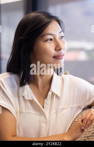 Im Büro sitzt lächelnde asiatische Frau auf der Couch, blickt weg und entspannt sich in der modernen Inneneinrichtung Stockfoto