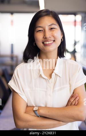 Lächelnde asiatische Frau, die mit gekreuzten Armen in einem modernen Büroumfeld steht Stockfoto