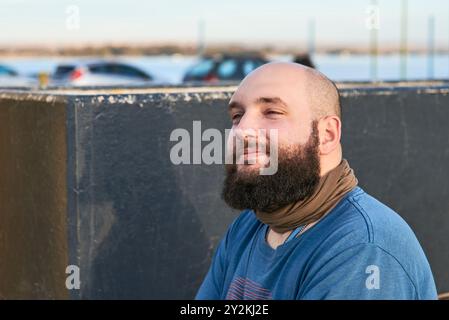 Porträt eines jungen bärtigen Mannes, der draußen sitzt und leicht lächelt, mit einem positiven, ruhigen und freudigen Ausdruck. Komposition mit Kopierraum. Stockfoto