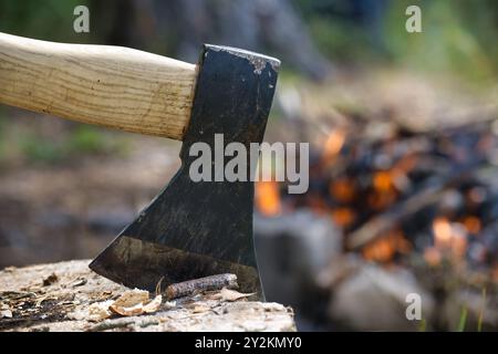 Nahaufnahme einer Holzfälleraxt, eingebettet in einen Baumstumpf, mit einem brennenden Lagerfeuer im Hintergrund während eines Campingausflugs. Stockfoto