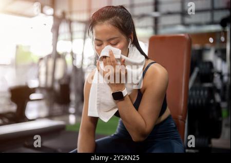 Eine müde, sportliche asiatische Frau in sportlicher Kleidung sitzt nach dem Workout im Fitnessstudio auf einer Bankpresse, um sich auszuruhen und mit einem Handtuch den Schweiß vom Gesicht abzuwischen. Arbeiten Stockfoto