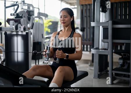 Eine schöne, sportliche asiatische Frau in sportlicher Kleidung sitzt auf einer Bank, zieht ein Seil, um ihre Muskeln im oberen Rücken zu trainieren, und trainiert auf einer Multistation Stockfoto