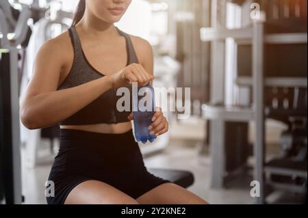 Ein zugeschnittenes Bild einer sportlichen, fitten asiatischen Frau in sportlicher Kleidung, die auf einer Bank sitzt, eine Wasserflasche öffnet und nach dem Training am G Wasser trinkt Stockfoto
