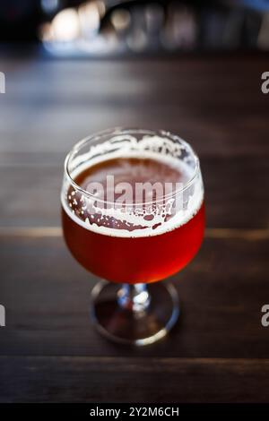 Ein Glas mit kräftigem Bernsteinbier steht auf einem dunklen Holztisch und fängt das warme Ambiente der gemütlichen Bar ein. Der schäumende Kopf klammert sich an Th Stockfoto