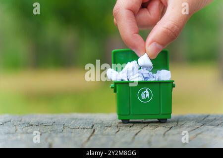 Nahaufnahme einer Hand, die zerknittertes Papier in einen grünen Miniaturmüll im Freien legt. Abfallbewirtschaftungs- und Recyclinggewohnheiten. Stockfoto