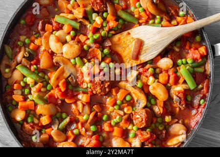 Spanische Chorizo, Butterbohnen, Tomaten- und Gemüseauflauf mit Pilzen und Zwiebeln. In einer Pfanne auf einem hölzernen Schneidebrett. Stockfoto