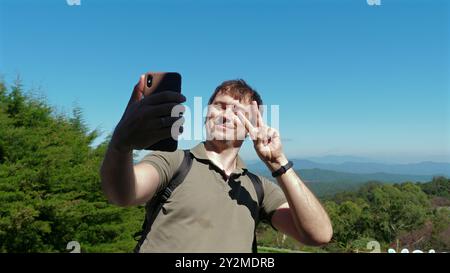 Ein Mann macht Selfie in der Natur, lächelt und zeigt Friedenszeichen, genießt Stockfoto