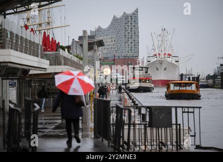 Hamburg, Deutschland. September 2024. Ein Passant mit Regenschirm läuft entlang der Landungsbrücken. Die markante Silhouette der Elbphilharmonie ist im Hintergrund neben dem Museumsschiff Cap San Diego zu sehen. Quelle: Christian Charisius/dpa/Alamy Live News Stockfoto
