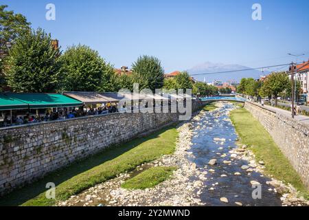 Tische und Stühle vor Bars, Cafés und Restaurants am Fluss Bistrica in der Altstadt von Prizren im Kosovo an einem sonnigen Tag. Stockfoto