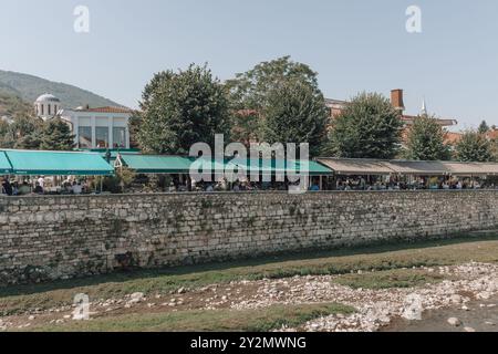 Tische und Stühle vor Bars, Cafés und Restaurants am Fluss Bistrica in der Altstadt von Prizren im Kosovo an einem sonnigen Tag. Stockfoto